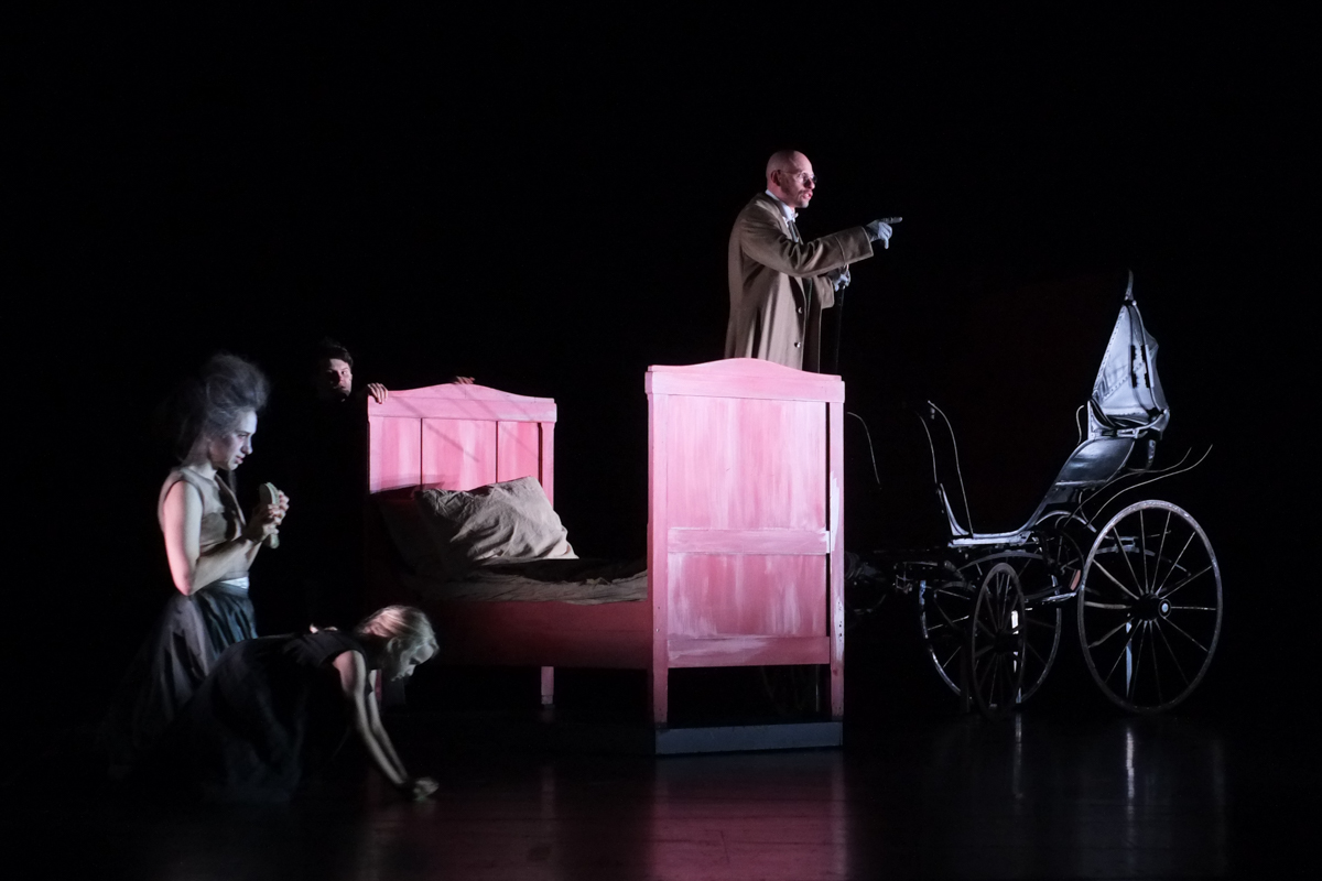 Pjotr Petrowitsch Luschin steht auf einem roten Bett, seine Moral verkündend, Polenka schrubbt den Boden, Rasumichin erscheint im Hintergrund aus dem Dunkel der Bühne am Schauspiel Stuttgart.