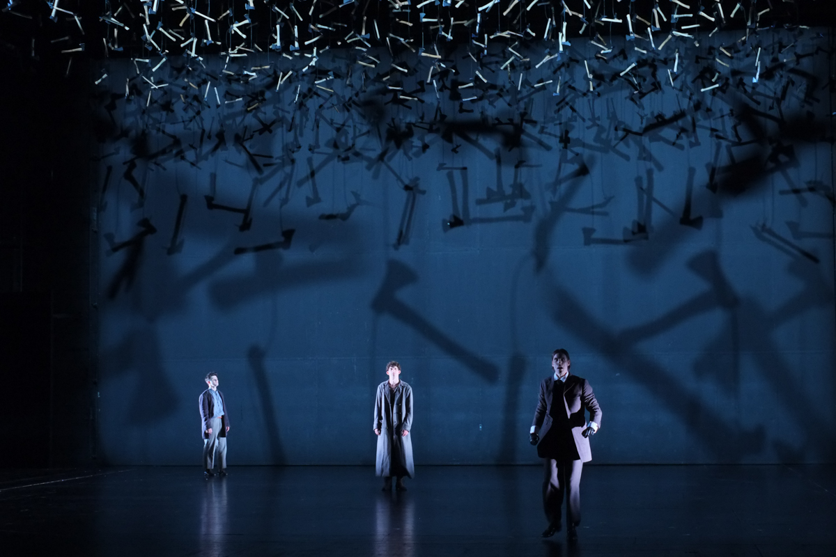 Szene am Schauspiel Stuttgart: Porfirij, Raskolnikow und Rasumichin auf der dunklen, scharf geleuchteten Bühne während sich ein Wald aus 3000 Äxten als Symbol der Schuld auf sie herabsenkt