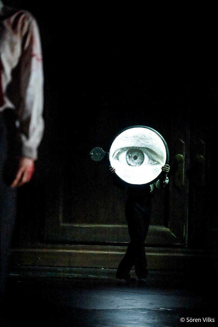 In a surreal 'Brott och Straff' scene at Dramaten Stockholm, Raskolnikov's bloody hand stands out in the foreground, while a giant watch, featuring a large eye instead of a dial, looms in the background, creating an enigmatic ambiance.